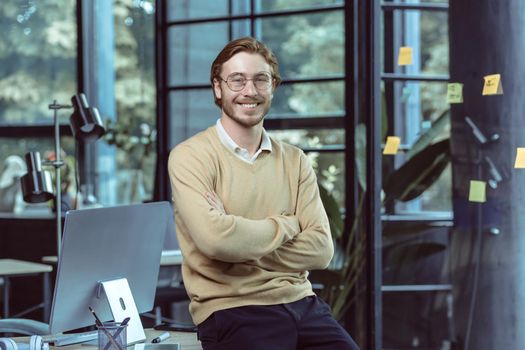 Portrait of successful young IT specialist young engineer programmer in modern loft office smiling and looking camera with crossed arms, businessman business owner wearing glasses and casual clothes