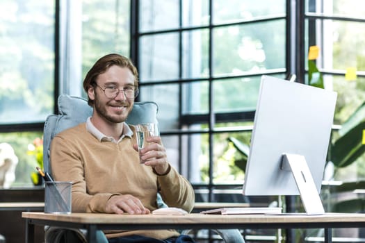 Young successful businessman man in modern office at work smiling and looking at camera, businessman holding glass of water, worker in glasses and casual clothes