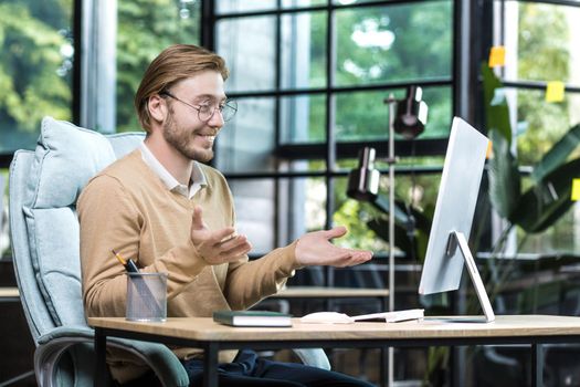 Happy and successful man at work looking at computer web camera smiling and talking with colleagues on video call, businessman working inside office building, remote online meeting with friends