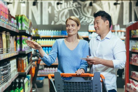 A young international couple, a woman and an Asian man walk between rows with a shopping cart in a supermarket, the woman points to the product, the man holds a shopping list, smilling.