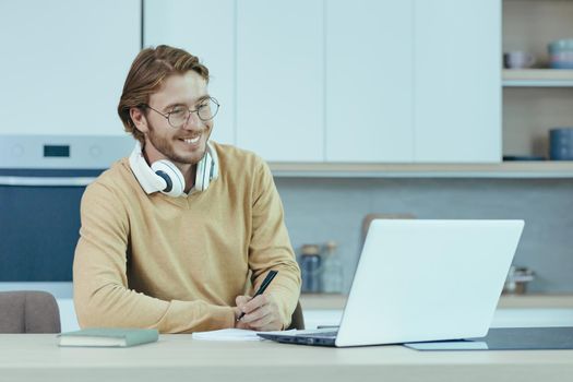 Young male entrepreneur working at home on paper work, sitting in the kitchen at the table, using a laptop for remote work as a freelancer.