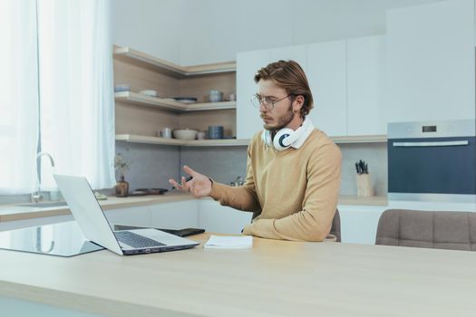 Young businessman working remotely from home sitting in kitchen, man using laptop for video call concentrating and talking seriously on online meeting with colleagues colleagues.