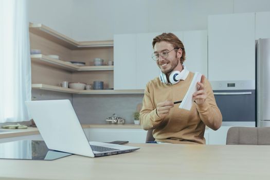 Young teacher teaches remotely from home, man works in kitchen at home, uses laptop for online call with communicates with students smiling explains assignment shows notebook.