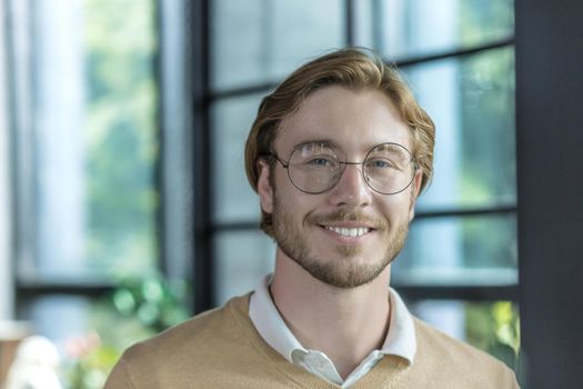 Portrait of a young handsome freelancer man, a blond businessman looking at the camera and smiling, a programmer in glasses works inside a modern office building, a close-up photo of a man at work