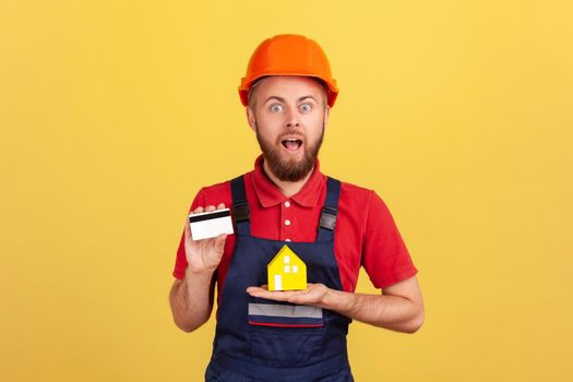 Portrait of shocked builder man wearing blue uniform and protective helmet showing credit card and paper house, looking at camera with open mouth. Indoor studio shot isolated on yellow background.