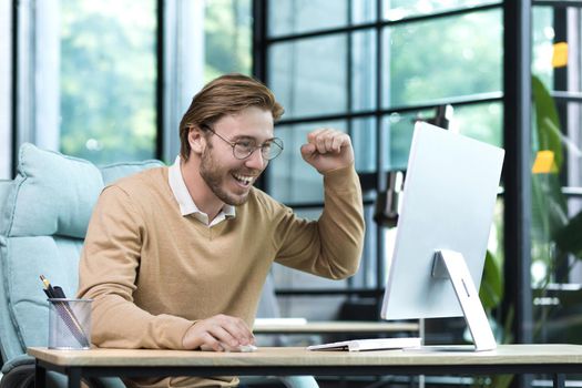 A successful young entrepreneur, a business owner, works in a modern office computer, a businessman , holds his hand up, celebrates victory and triumph received good joyful news result