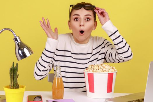 Portrait of shocked impressed woman sitting at workplace with popcorn and beverage, raising glasses and watching shocking moment. Indoor studio studio shot isolated on yellow background.