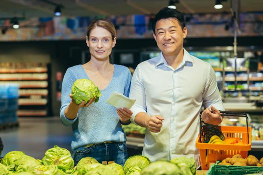 Portrait of a young interracical couple, a European woman and an Asian man buy vegetables, green cabbage in a supermarket according to a list. They look at the camera, smile.