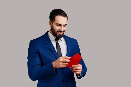 Handsome bearded businessman pulling letter from red envelope, holding greeting card and smiling joyfully, wearing official style suit. Indoor studio shot isolated on gray background.