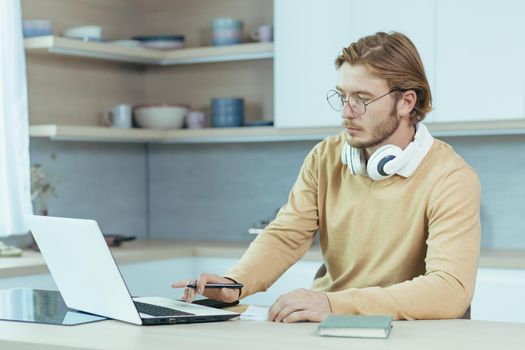 Young serious and focused man studying remotely at home, student in kitchen wearing glasses using laptop for distance learning taking notes on study material.
