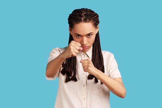 Woman with black dreadlocks having serious and confident look, keeping fists clenched in front of her, defencing herself from offence and bad treatment. Indoor studio shot isolated on blue background.