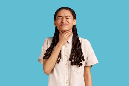Unhappy woman with dreadlocks has terrible throat pain, suffers from suffocation, touches neck, frowns face, difficult to swallow, wearing white shirt. Indoor studio shot isolated on blue background.