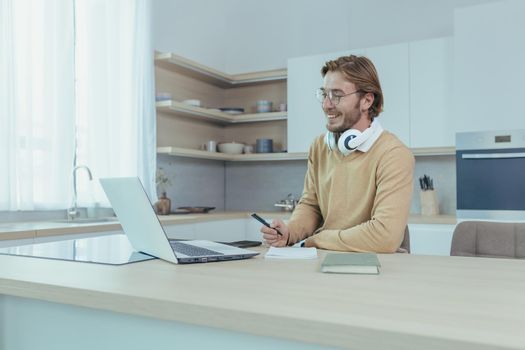 Young successful and happy businessman working from home, man using laptop for remote work, freelancer smiling in casual clothes sitting in kitchen at table.