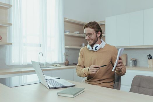 Young teacher teaches remotely from home, man works in kitchen at home, uses laptop for online call with communicates with students smiling explains assignment shows notebook.