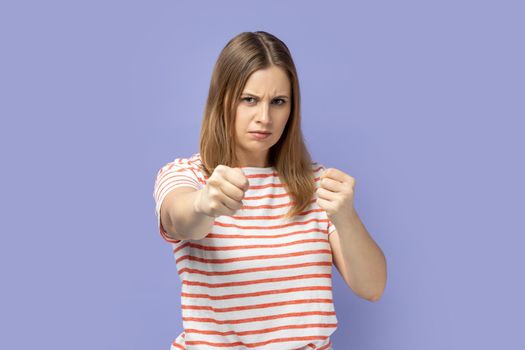 Portrait of aggressive blond woman wearing striped T-shirt holding clenched fists up, fighting with abuser, defence from domestic violence. Indoor studio shot isolated on purple background.