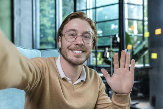 Man working in modern office, happy blond businessman smiling and looking at smartphone camera, holding hand up greeting gesture, worker in glasses taking selfie, video call with friends