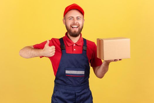 Cargo transportation, delivery service. Smiling courier man in uniform holding cardboard box and showing thumb up, delivering parcel to client. Indoor studio shot isolated on yellow background.