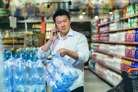 A worried young Asian man buys pallets of water, many bottles of water in a supermarket, shop. Talking on the phone. Panic during quarantine, corona virus, covid.