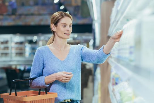 A young beautiful housewife is standing in a supermarket with a basket in her hand, choosing dairy products. He takes a bottle with milk, yogurt, smiles.