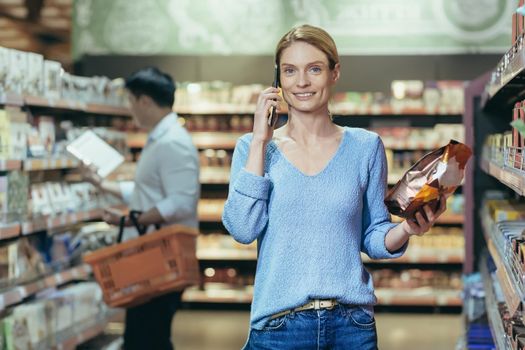 A beautiful young blonde woman is standing in a supermarket and talking on the phone, consulting, holding a pack of coffee in her hand, smiling. Shoppers in the background.
