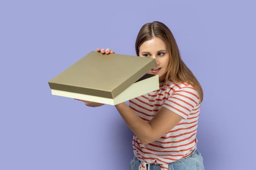 Portrait of curious calm blond woman wearing striped T-shirt standing holding present box and looking inside, interesting what inside. Indoor studio shot isolated on purple background.