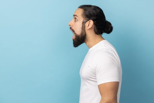 Side view of surprised amazed man with beard wearing white T-shirt looking with shocked expression away, sees something astonishing. Indoor studio shot isolated on blue background.