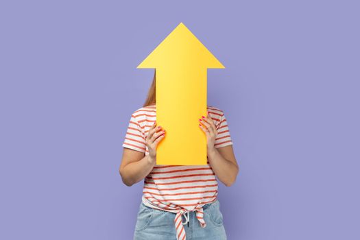 Portrait of anonymous unknown blond woman wearing striped T-shirt holding big yellow arrow pointing up, showing increase in career. Indoor studio shot isolated on purple background.