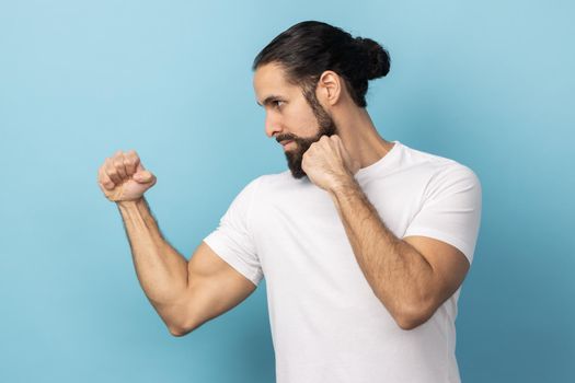 Side view of aggressive man with beard wearing white T-shirt holding clenched fists up ready to boxing, martial art trainer, self defense. Indoor studio shot isolated on blue background.