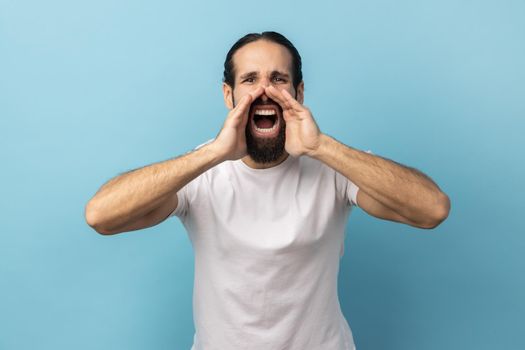 Attention, announcement. Portrait of handsome man with beard wearing white T-shirt screaming message, loud advertising, keeps hands near mouth. Indoor studio shot isolated on blue background.