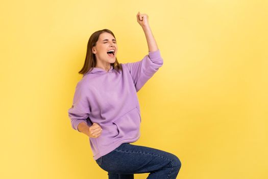 Portrait of enthusiastic woman screaming for joy and raising high her hand, celebrating successful winning, incredible victory, wearing purple hoodie. Indoor studio shot isolated on yellow background.