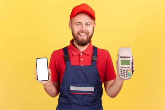 Portrait of smiling delighted bearded worker man standing and holding pos terminal and smartphone with blank screen for your advertisement. Indoor studio shot isolated on yellow background.