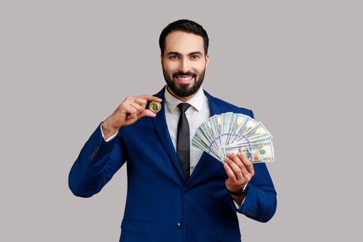 Man holding in hands and showing dollar bills and golden coin of crypto currency, transferring money into digital bitcoins, wearing official style suit. Indoor studio shot isolated on gray background.