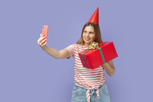 Portrait of blond woman blogger wearing striped T-shirt and party cone holding present box and making selfie or having video call. Indoor studio shot isolated on purple background.