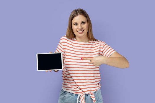 Portrait of smiling blond woman wearing striped T-shirt pointing at tablet empty screen for adv, looking at camera with toothy smile. Indoor studio shot isolated on purple background.