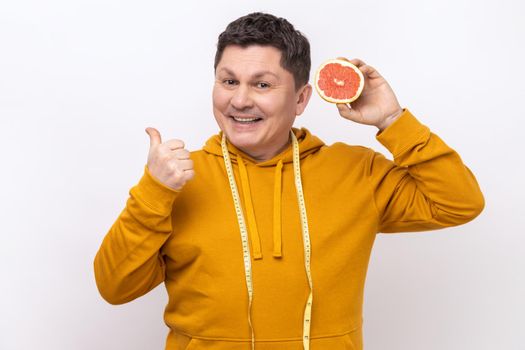 Middle aged man with dark hair holding slice of grapefruit, looking at camera with toothy smile, showing thumb up, wearing urban style hoodie. Indoor studio shot isolated on white background.