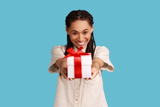 Portrait of happy woman with black dreadlocks giving present box, looking at camera, congratulating friend with birthday, wearing white shirt. Indoor studio shot isolated on blue background.