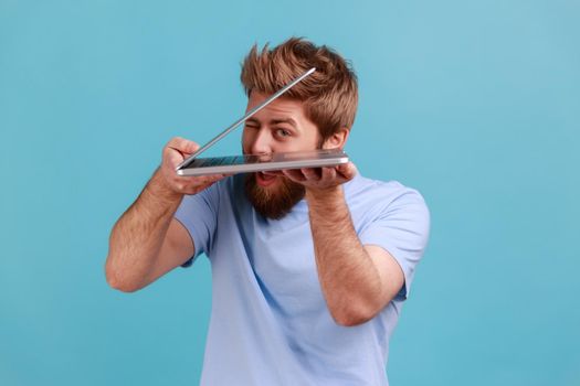 Portrait of handsome young adult bearded man taking a look at laptop display, trying to see private information, spying via web camera. Indoor studio shot isolated on blue background.