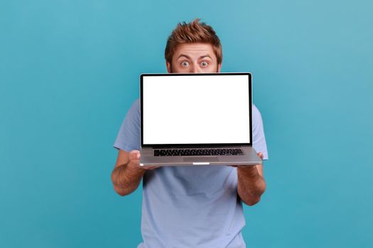 Portrait of man hiding behind white empty screen laptop, surprised with advertising area, looking at camera with big eyes, updating operation system. Indoor studio shot isolated on blue background.