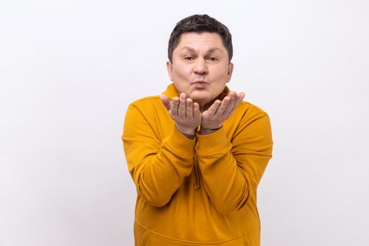 Love you. Handsome man sending air kiss over palms, flirting to camera, demonstrating romantic amorous feelings, wearing urban style hoodie. Indoor studio shot isolated on white background.