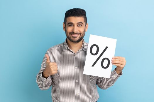 Smiling smart businessman showing paper with percent sign inscription, looking at camera, showing thumb up, wearing striped shirt. Indoor studio shot isolated on blue background.