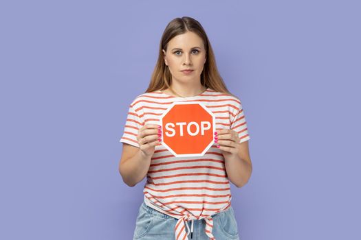 Portrait of serious beautiful blond woman wearing striped T-shirt holding red stop sign and looking at camera with dissatisfied expression. Indoor studio shot isolated on purple background.