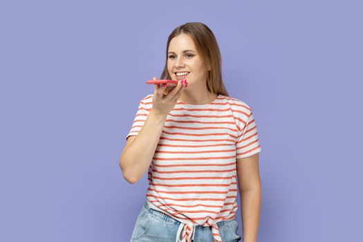 Portrait of smiling delighted positive blond woman wearing striped T-shirt standing with smart phone and recording voice message, voice assistant. Indoor studio shot isolated on purple background.