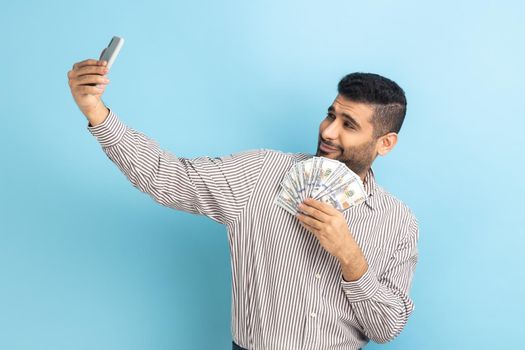 Extremely satisfied businessman making selfie photo with bunch of dollars on smartphone, bragging with wealth, wearing striped shirt. Indoor studio shot isolated on blue background.