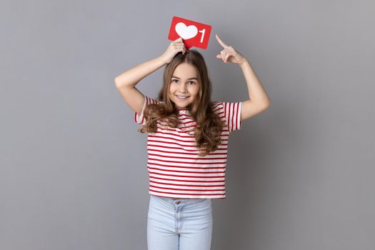 Portrait of happy little girl wearing striped T-shirt holding above head and pointing at heart like icon, recommending to click on social media button. Indoor studio shot isolated on gray background.