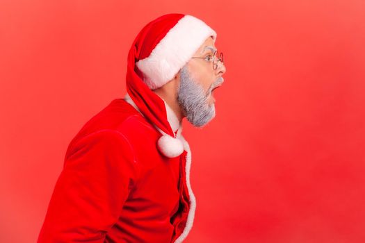 Side view portrait of elderly man with gray beard wearing santa claus costume looking ahead with opened mouth, hearing shocking news. Indoor studio shot isolated on red background.