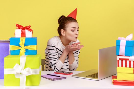 Charming woman in party cone sending air kisses to laptop display, sitting at workplace, receive congratulations from friends by video call. Indoor studio studio shot isolated on yellow background.