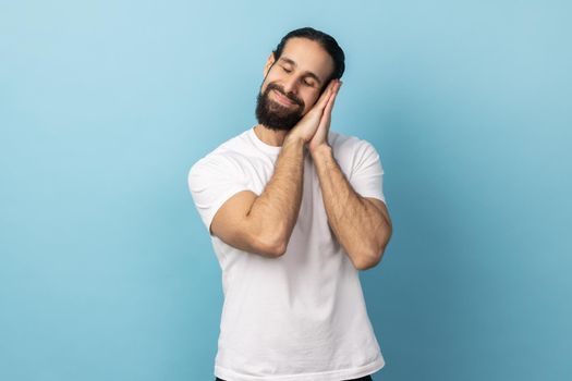 Portrait of tired exhausted handsome man with beard wearing white T-shirt sleeping laying down on her palms, keeping eyes closed. Indoor studio shot isolated on blue background.