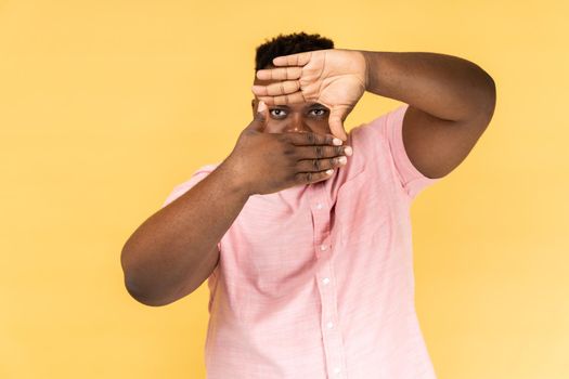 Capture moments. Portrait of man wearing pink shirt looking through photo frame shape with fingers, viewing distant, focusing and zooming at camera. Indoor studio shot isolated on yellow background.