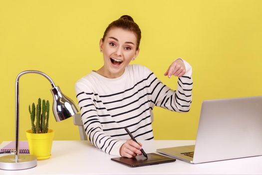 Delighted woman creative designer sitting at workplace with graphic tablet and laptop, pointing at camera with excitement. Indoor studio studio shot isolated on yellow background.