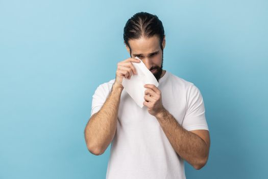 Portrait of sad upset handsome man with beard wearing white T-shirt standing, holding his head down and crying, having serious problems. Indoor studio shot isolated on blue background.